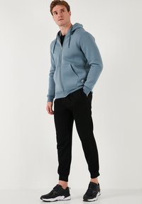 Sudadera con cremallera de color azul claro, con capucha y bolsillos frontales, combinada con pantalones negros ajustados y zapatillas grises. Textura de tela suave.