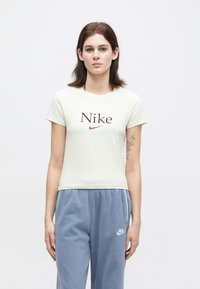 Mladá žena s krátkymi hnedými vlasmi, oblečená v bielom tričku s logom Nike a modrých teplákoch Nike, stojí pred jednobarevným bielym pozadím.