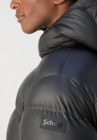 Zwarte gewatteerde jas met een glanzende, textuur afgewerkte finish, voorzien van een capuchon en een zichtbare logo patch op de schouder. Accenten met een strakke vormgeving.