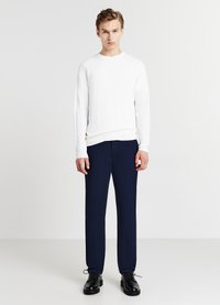 Weißer gerippter Strickpullover mit einem runden Ausschnitt, kombiniert mit dunkelblauen Hosen und schwarzen Schnürschuhen. Glatte Textur, figurbetontes Design.