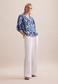 Bluse mit blauem Blumenmuster, V-Ausschnitt und kurzen Ärmeln, kombiniert mit weißen weit geschnittenen Hosen und schwarzen Sandalen. Glatte Stofftextur.