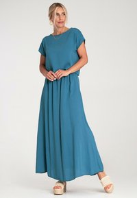 Maxi sukienka w turkusowym materiale, posiada krótkie rękawy, luźny krój, marszczoną talię i sięga do kostek. Noszona z sandałami na platformie z paskami.