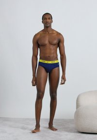 Μοντέλο που φοράει εσώρουχα σε σκούρο μπλε χρώμα με φωτεινή κίτρινη ζώνη μέσης που φέρει το λογότυπο "Nike". Το δέρμα είναι γραμμωμένο και το φόντο είναι ανοιχτό γκρι.