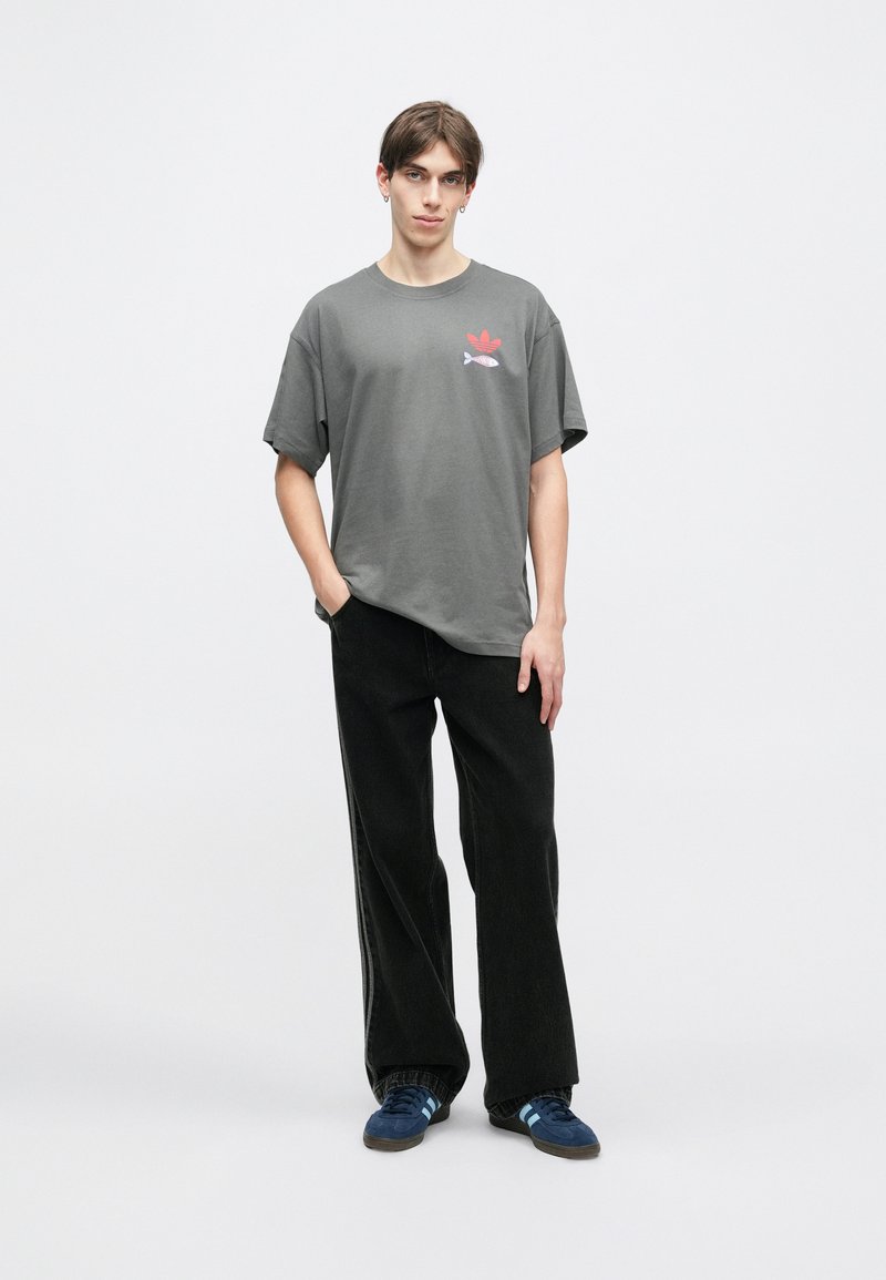 Camiseta de algodón gris con mangas cortas y un estampado gráfico de elementos rojos y azules. Combinada con pantalones anchos negros y zapatos azules.