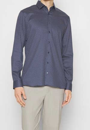 Mann trägt ein marineblaues langärmeliges Hemd mit weißen Punkten und helle beige Hose, steht vor einem einfarbigen Hintergrund.