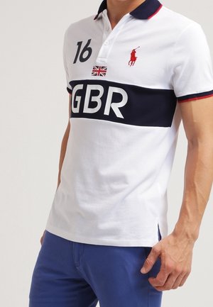 Mand iført hvid polo med en marineblå stribe, "GBR" bogstaver, nummer 16, britisk flag og rød hestelogo, kombineret med blå bukser.
