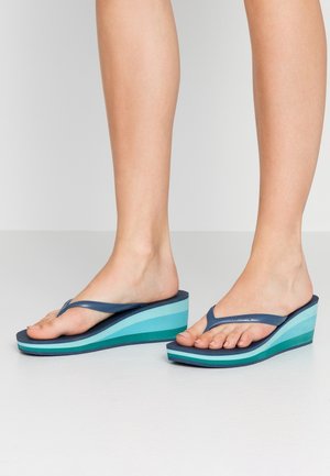 Fødder iført kilehæl-sandaler med blå gradient-såler og marineblå stropper, stående på en glat, lysegrå gulv.
