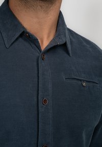 Navyblauwe overhemd met een gestructureerde weving, knoopsluiting aan de voorkant en een enkel borstzakje. Heeft bruine knopen en een puntige kraag.