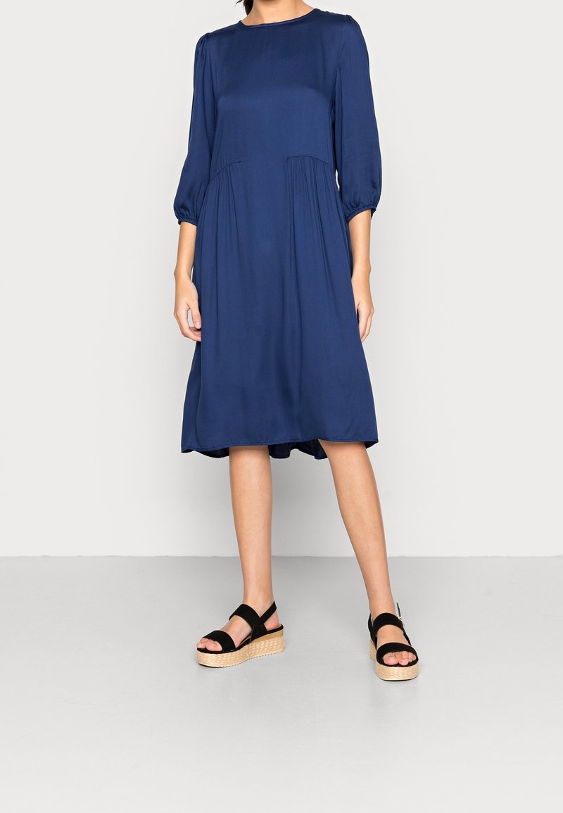 Femme portant une robe bleu marine longueur genou avec des manches trois quarts et des sandales compensées noires à semelles tressées.