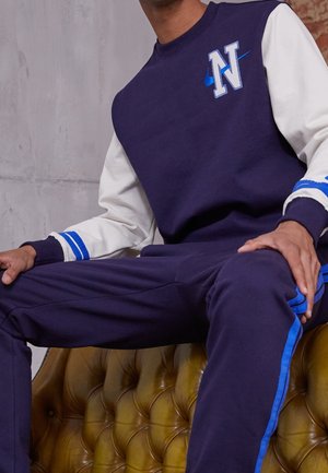 Mann trägt marineblaue und weiße Sweatshirt mit blauem "N" und Nike Swoosh-Logo, sitzt auf einem braunen, gesteppten Ledersessel.