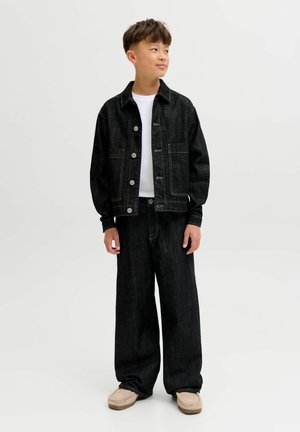 Poiss seisab küljele vaadates, seljas must teksajakk ja laia säärega teksad, valge särgi ning beežide jalanõudega.