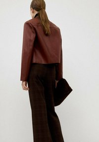 Giacca cropped in pelle marrone con una texture liscia, indossata sopra pantaloni a quadri scuri. Accessorio pochette nera tenuto in mano, design minimalista.