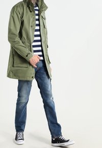 Olivově zelená bunda s kapucí, pod ní modrobíle pruhované tričko, modré džíny a černé tenisky s bílými detaily.