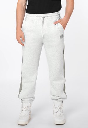 Man draagt lichtgrijze joggingbroek met zijstrepen en witte sneakers, handen gedeeltelijk in de zakken, staand tegen een witte achtergrond.