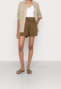 Shorts vert olive avec ceinture élastique, chemise à carreaux beige, et sandales vertes à enfiler. T-shirt blanc à col en V en dessous.