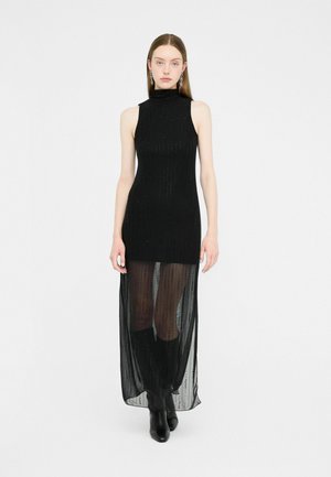 Black sleeveless dress with a high neckline, sheer long hem, textured fabric, and vertical stripe embellishments. Worn with knee-high boots.