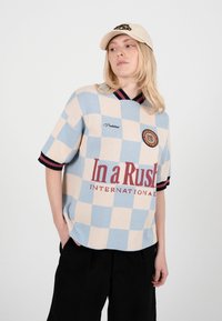 Ung person med langt blondt hår iført en beige kasket, blå og beige ternet skjorte med teksten "In a Rush" og sorte bukser mod en hvid baggrund.