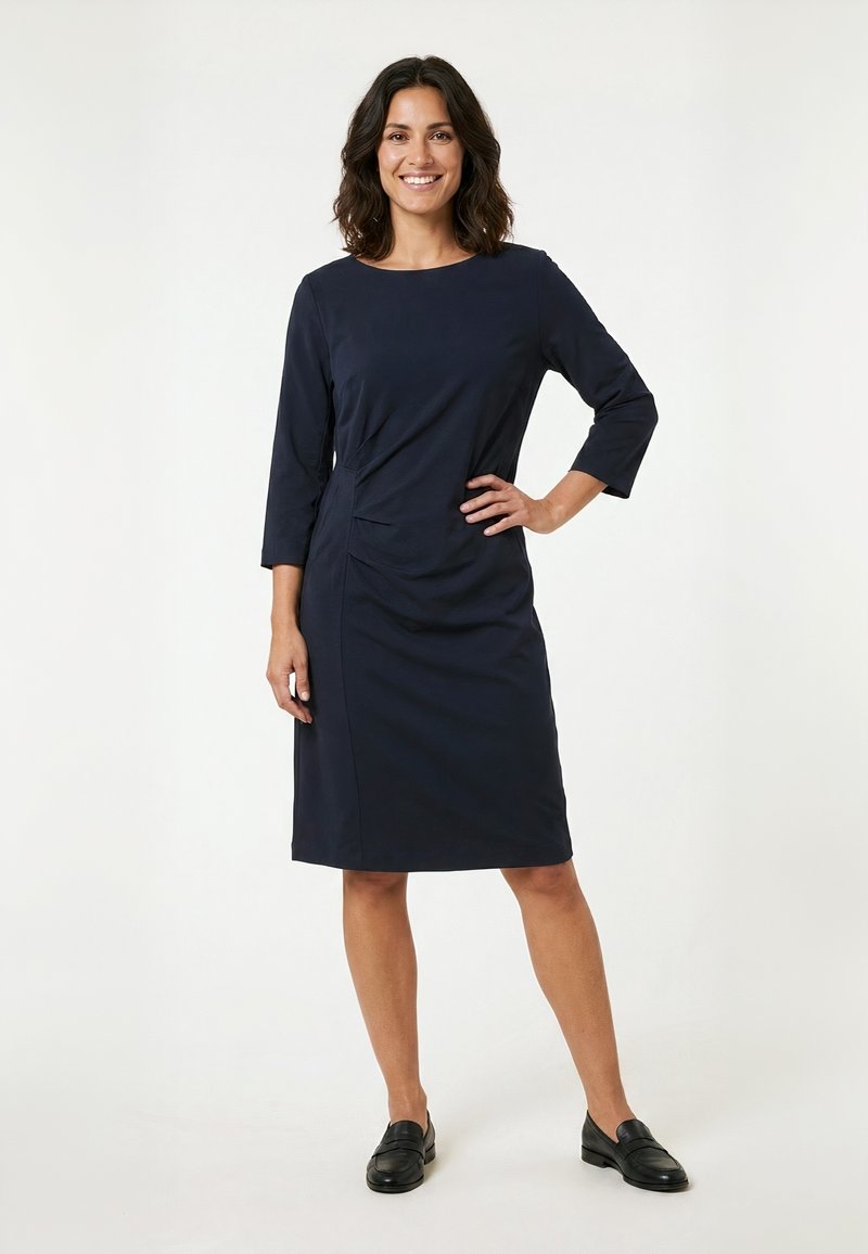 Femme souriante aux cheveux foncés mi-longs, portant une robe bleu marine longueur genoux avec des manches trois-quarts et des mocassins noirs, debout avec la main sur la hanche.