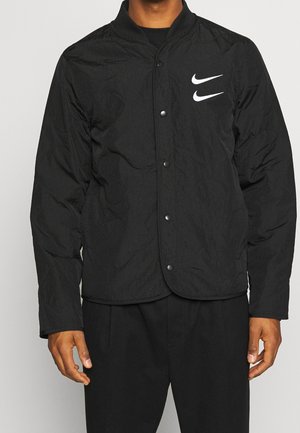 Homme portant une veste noire à boutons avec deux logos Nike swoosh blancs sur le côté gauche de la poitrine, associée à un pantalon noir.