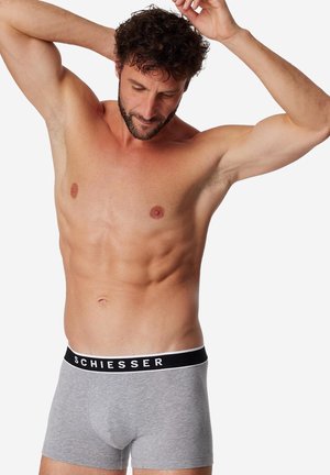 Borstloze man met donker krullend haar, gekleed in lichtgrijze boxershorts met "SCHIESSER" op de zwarte tailleband, poseert met opgetilde armen.