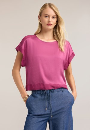 Blonde Frau mit langen Haaren, die goldene Ohrringe und Armbänder trägt, ein magentafarbenes kurzärmeliges Oberteil und blaue Hose mit Kordelzug, Hände in den Taschen.