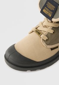 Sapatilhas Palladium em bege e olive com biqueira em borracha preta, atacadores com "Hopper" e logótipo de "Stranger Things" na língua.