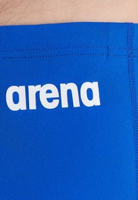 Bañador azul hecho de una tela suave con el texto de marca blanco "arena". La costura es visible a lo largo de los bordes para mayor durabilidad.