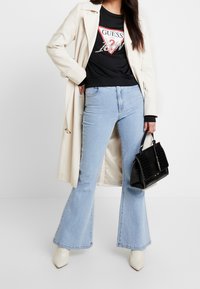 T-shirt nera GUESS con logo, trench crema, jeans svasati blu chiaro e piccola borsa nera in pelle cocco. Tacchi a punta bianchi.
