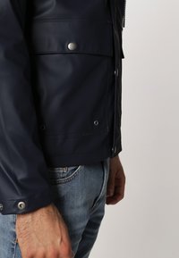 Marinblå vattentät jacka med en slät yta, tryckknappar och en ficka fram. Designen har en åtsittande passform och en lätt glänsande yta.