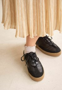 Musta lenkkikenkä, jossa yhdistyy sileä ja teksturoitu materiaali, pyöreät nauhat ja konjakinvärinen kumipohja, käytetty valkoisten röyhelöisten sukkien kanssa.