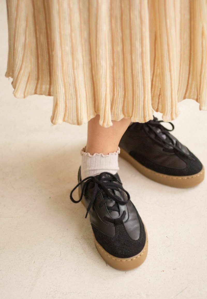 Musta lenkkikenkä, jossa yhdistyy sileä ja teksturoitu materiaali, pyöreät nauhat ja konjakinvärinen kumipohja, käytetty valkoisten röyhelöisten sukkien kanssa.