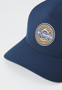 Námornícky modrá čiapka s plochým okrajom, textúra látky a okrúhly logo Columbia Sportswear Company so zlatými a bielymi horami a stromami.
