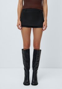 Fekete, magas derekú mini szoknya sima felülettel és diszkrét oldalsó zsebekkel, térdig érő fekete bőr csizmával párosítva.