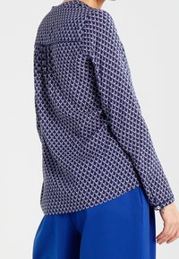 Blusa a fantasia navale con design geometrico bianco, maniche lunghe, scollatura rotonda e dettaglio a pieghe sul retro, indossata con pantaloni blu.
