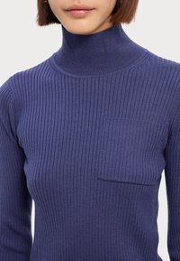 Navyblauer, gerippter Rollkragenpullover mit langen Ärmeln und einer kleinen Fronttasche. Der Stoff scheint weich und enganliegend zu sein.