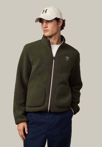 Oliivinvihreä fleece-takki, jossa vetoketju edessä, ruskeat yksityiskohdat ja kaksi isoa taskua, yhdistetty valkoiseen lippalakkiin, jossa tumma "H."