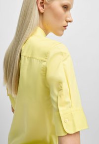 Gelbes Hemd mit Kragen aus glattem, leichtem Stoff. Verfügbar mit hochgerollten Ärmeln und Knopfdetails sowie einem einfachen, klaren Design.