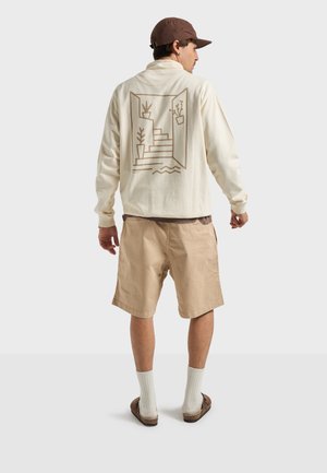 Man draagt een bruine pet, een crèmekleurig shirt met lange mouwen met een ontwerp van een trap en planten op de achterkant, beige korte broek, witte sokken en bruine sandalen.