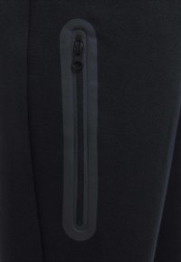 Un pantalon de sport noir présente une poche zippée avec un design élégant et une texture lisse, améliorant la fonctionnalité et le style.