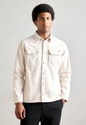 Light beige button-up shirt made of cotton, featuring two chest pockets, long sleeves, and a collared neckline, paired with black pants.