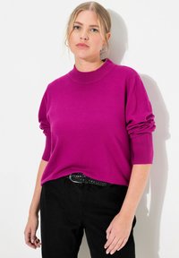 Fuchsový pletený sveter s vysokým golierom a vyrolovanými rukávmi, spojený s čiernymi nohavicami s vysokým pásom a pleteným čiernym opaskom. Hladká textúra.