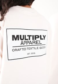 Sudadera de color rosa claro con una gran etiqueta blanca en la parte posterior que lleva texto en negro: "MULTIPLY APPAREL", "CRAFTED TEXTILE GOODS" y "EST. 2015."