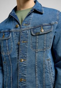 Chaqueta de denim azul oscuro con un diseño clásico, que presenta botones de latón, bolsillos en el pecho y detalles de costura sutiles. Textura suave, ajuste casual.