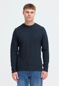Maglietta a maniche lunghe blu navy con tessuto texturizzato, scollo rotondo e un piccolo logo sul petto, abbinata a jeans azzurri chiari.