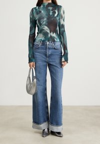 Long-sleeve, ribbed top in teal with dark patterns, paired with high-waisted, wide-leg denim jeans with light blue cuffs and silver handbag.