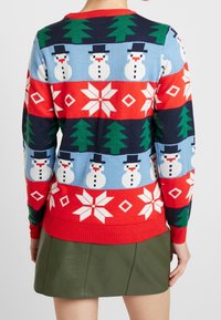 Stickad tröja med flerfärgade snögubbar, träd och geometriska mönster. Har en röd halsringning och ribbad nederkant. Bärs över en grön kjol.