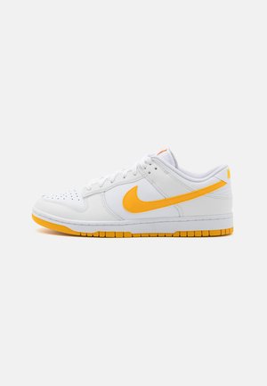 White Nike sneakers with a smooth leather finish, featuring a bold orange swoosh, textured sole, and contrasting yellow accents.