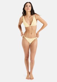 Geel bikini met bloemenpatroon; bralette-stijl top met diepe V-hals en decoratieve knop; verstelbare strik-rokken onderkant. Glad stof textuur.