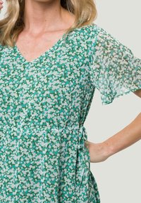 Vestido verde y azul claro con escote en V, mangas cortas transparentes y un detalle de cintura con lazo. Presenta una tela texturizada con un diseño estampado.