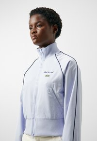 Chaqueta de cremallera de color púrpura claro en tejido transpirable, con cuello y puños acanalados, logo bordado y ribete lateral en contraste.